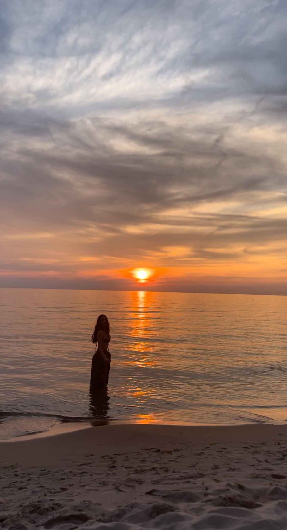 The beach at sunset, an orange and purple sky and a girl's silhouette in the water looking at the camera.