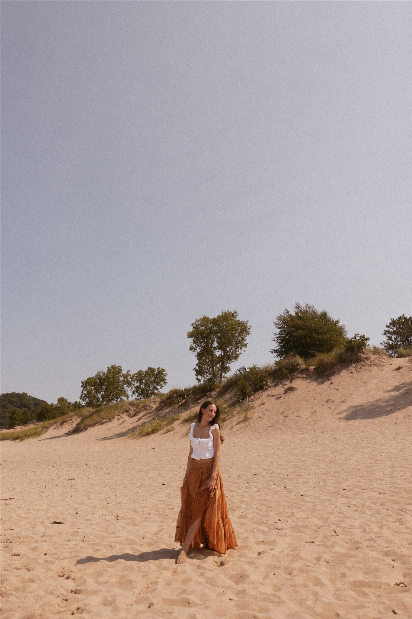 Girl standing and smiling on beach in terracotta colored skirt and white top.