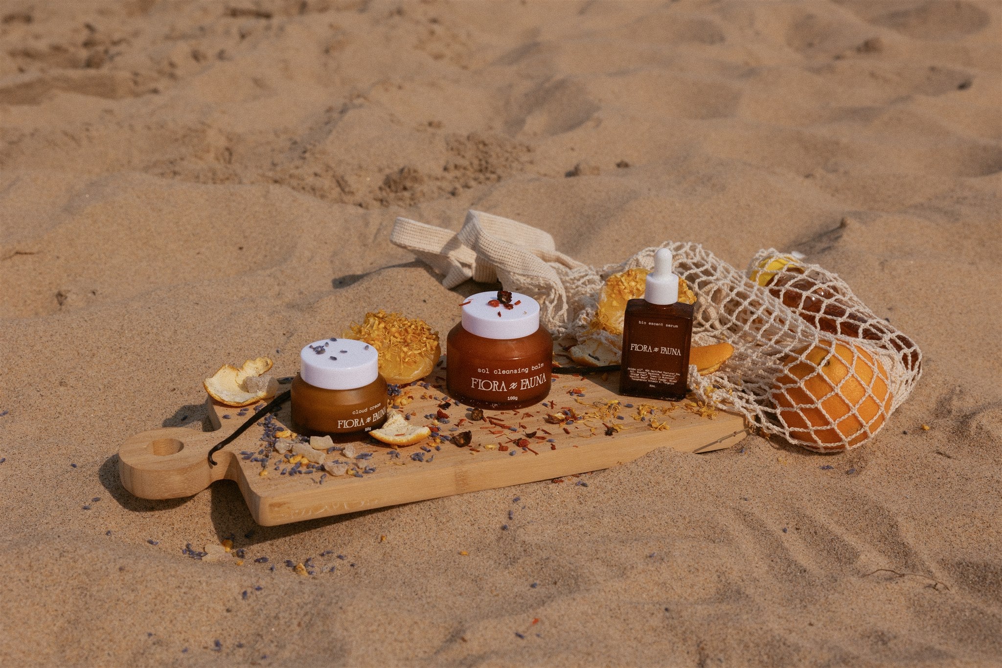 Jar of cream, balm, and bottle of serum displayed on wooden serving board in the sand, with oranges, lavender, and vanilla. 