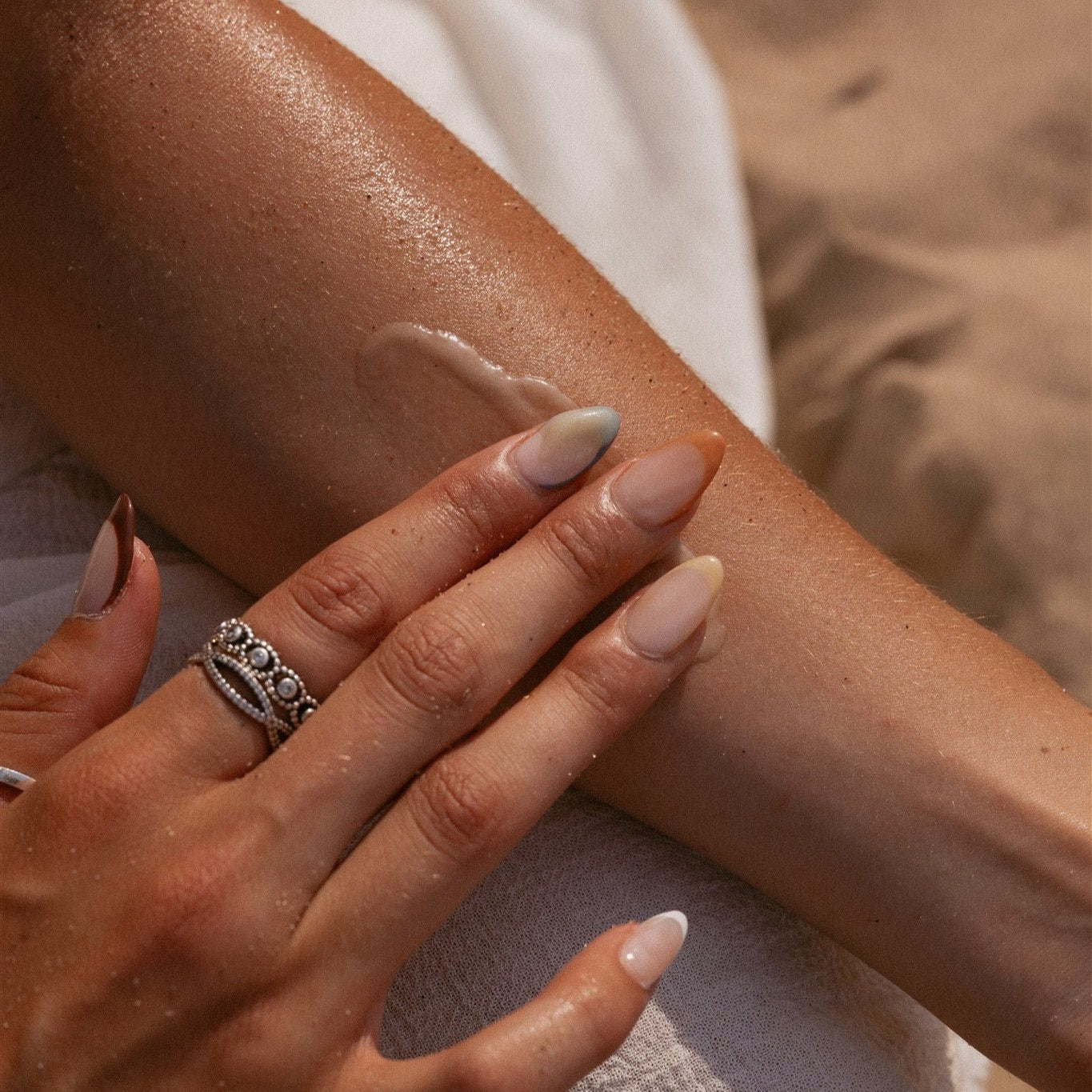 A girl's hand with a silver ring on her index finger and tan skin applying cream to the inside of her arm at the beach. 