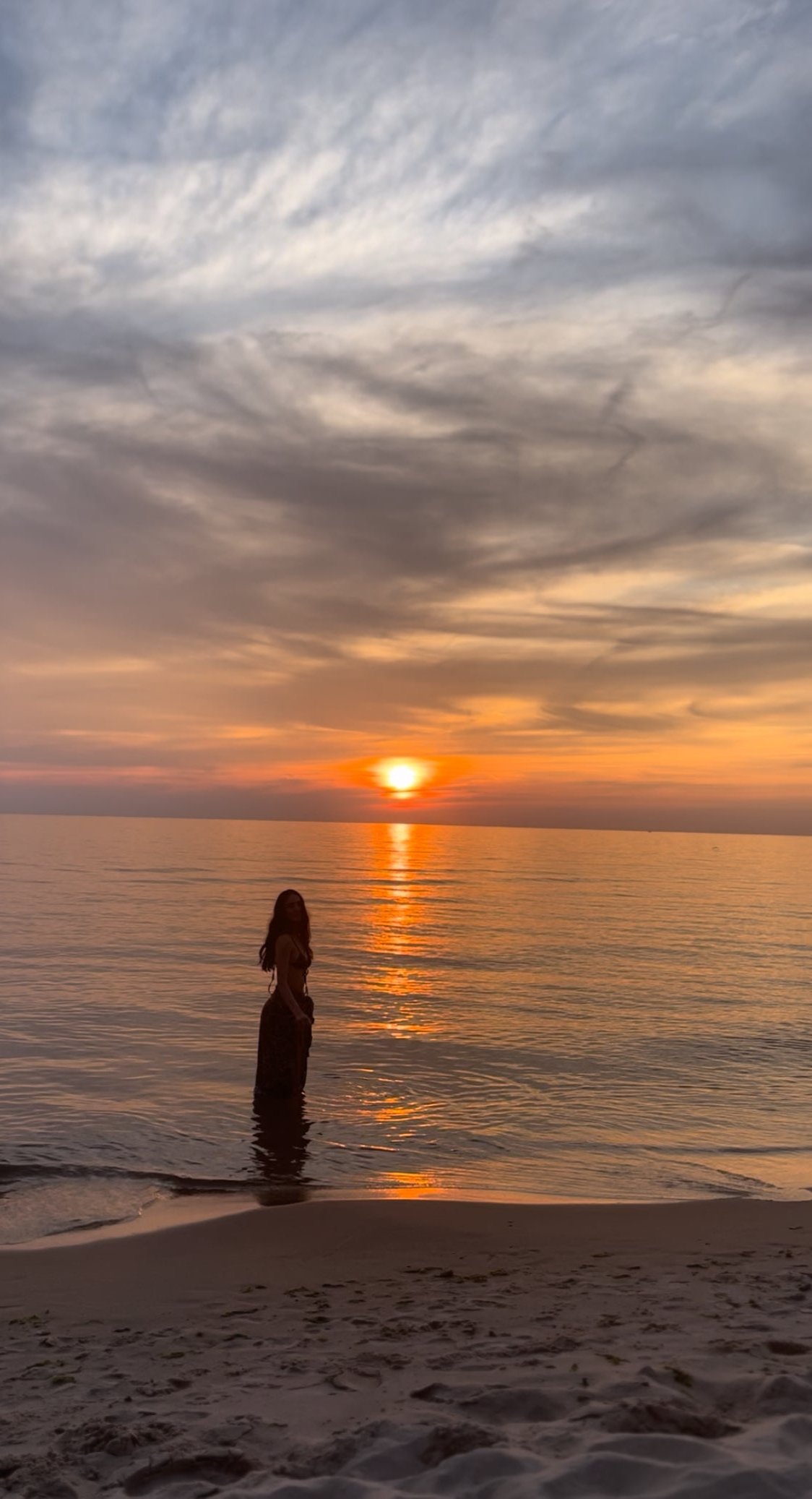 The beach at sunset, an orange and purple sky and a girl's silhouette in the water looking at the camera. 