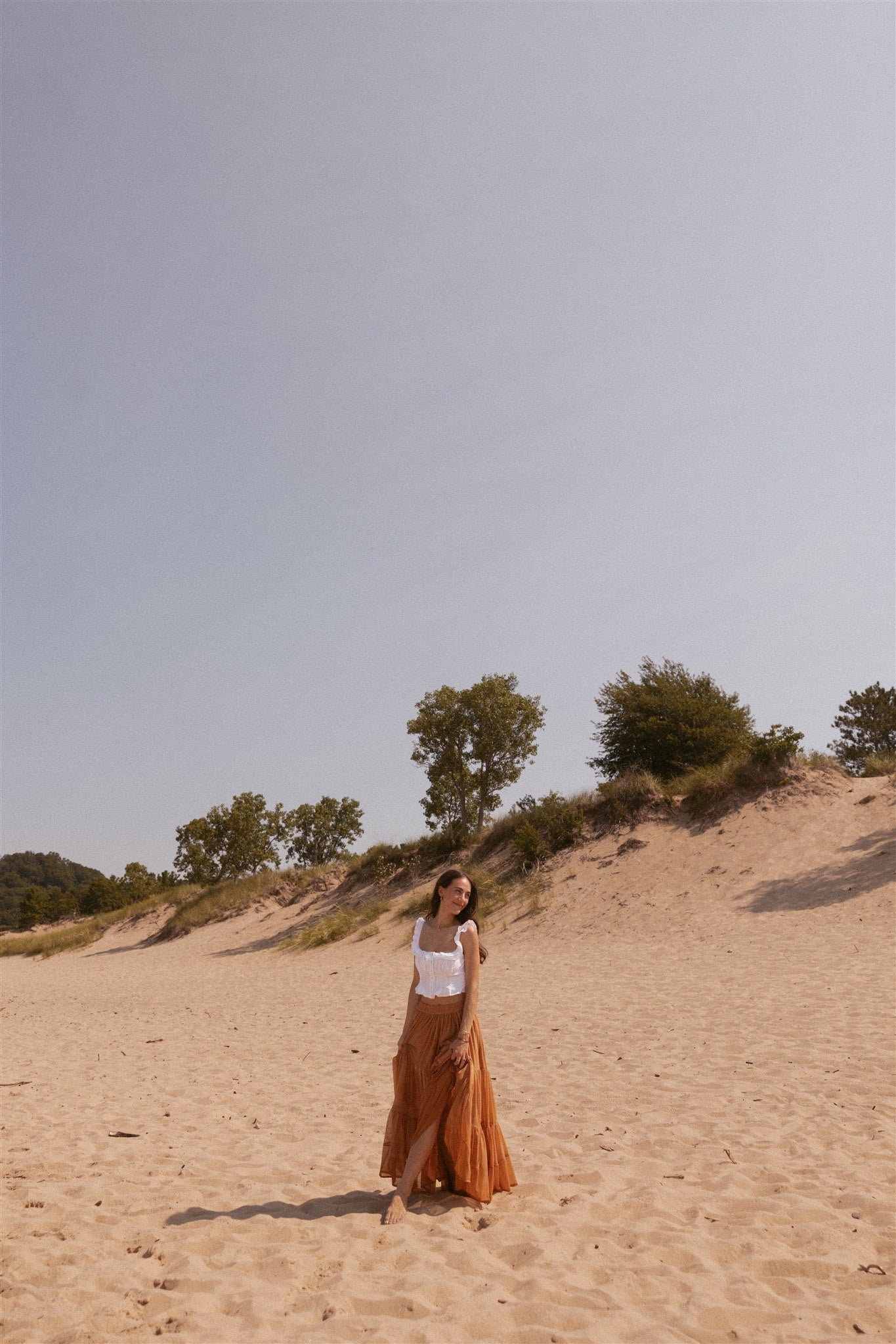 Girl standing and smiling on beach in terracotta colored skirt and white top. 