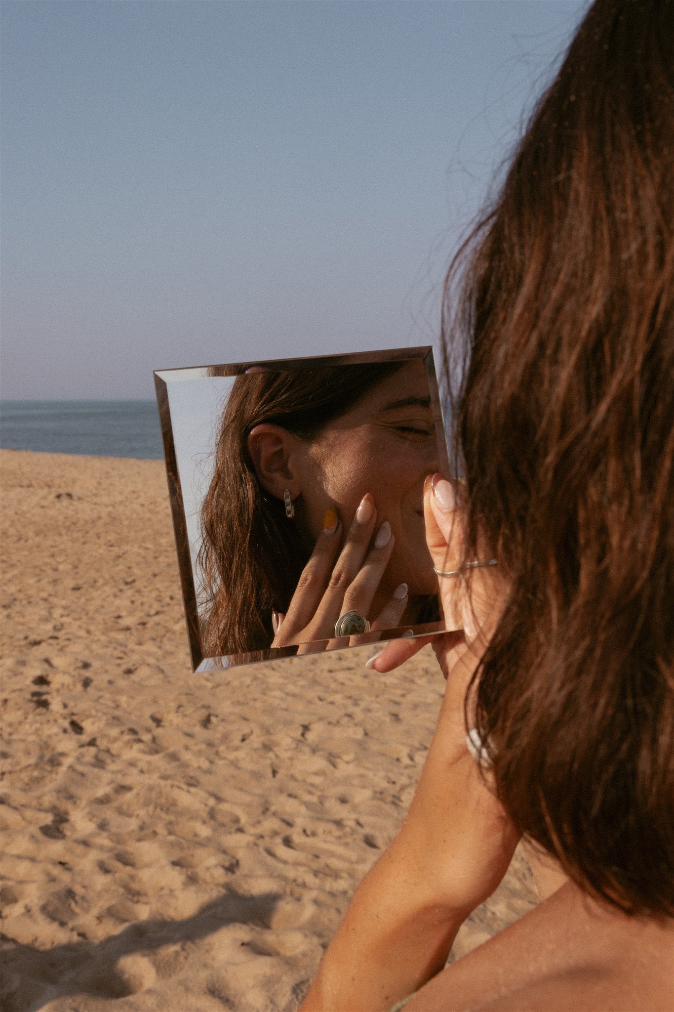 A girl looking into a square mirror at the beach, applying the orange sol balm with fingers.