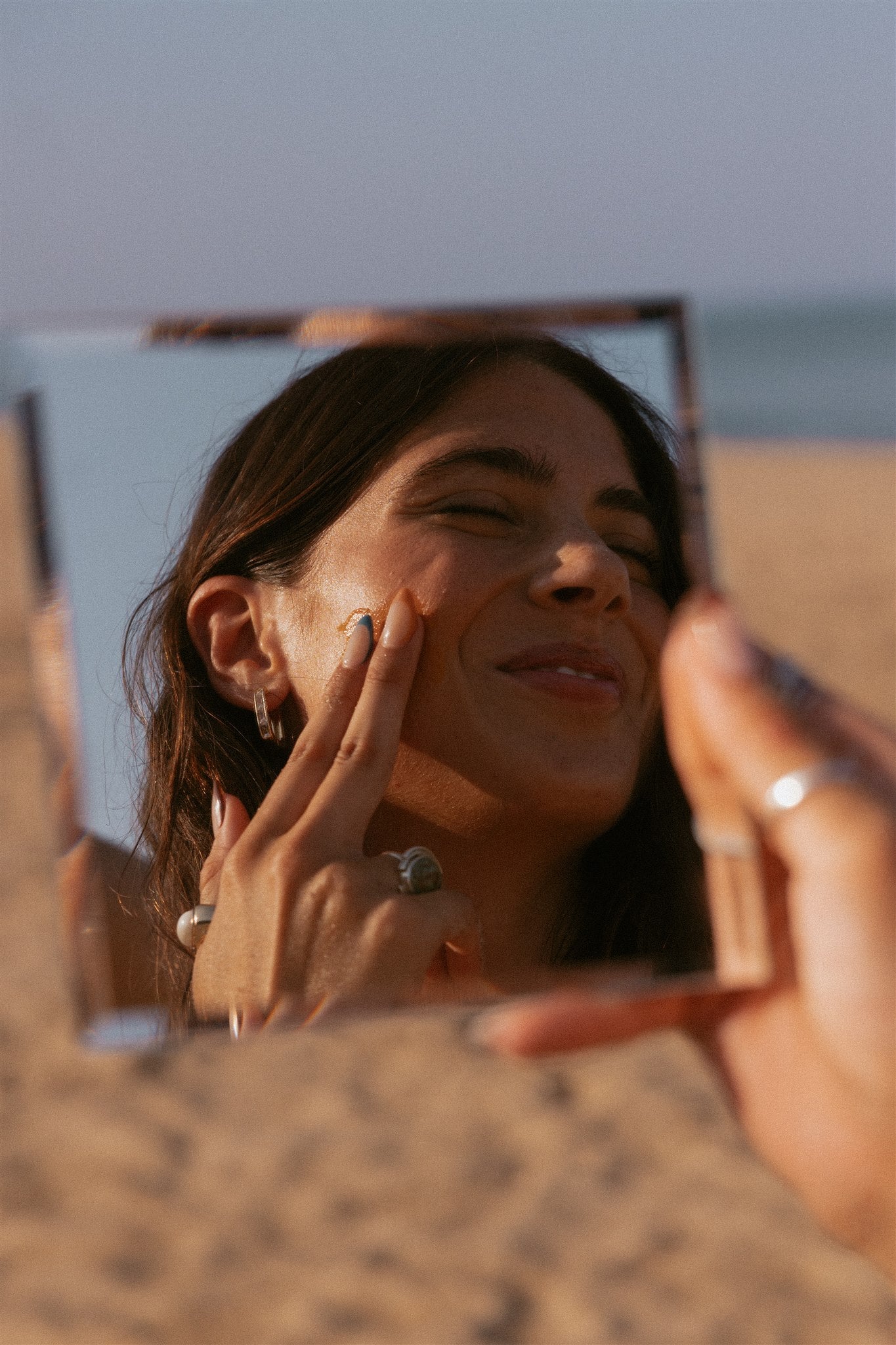 Smiling girl applying cleansing balm to face, using a mirror on the beach. 